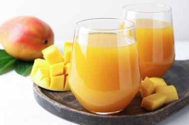 Bardakta lezzetli mango suyu, taze meyve ve yeşil yapraklar beyaz masada, yakın plan.