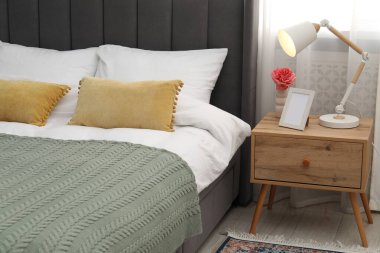 Yatağın yanında lamba, çiçek ve çerçeve olan komodin.