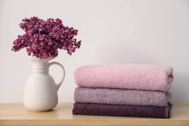 Beyaz duvarın yanındaki ahşap masada yumuşak havlular ve leylak çiçekleri yığını.