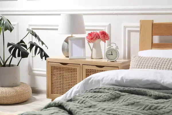 İçinde lamba, çalar saat ve yatağın yanında çiçekler olan komodin.