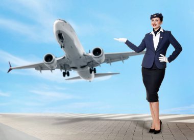 Gülümseyen hostes bulutlu mavi gökyüzünü gösteren uçağı işaret ediyor.