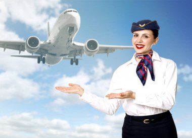 Gülümseyen hostes bulutlu mavi gökyüzünü gösteren uçağı işaret ediyor.