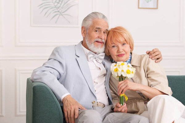 Lovely senior couple with bouquet of flowers on sofa at home