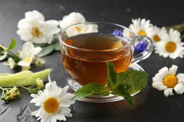 Cam fincanda aromatik bitki çayı, farklı çiçekler ve siyah masada nane şekeri, yakın plan.