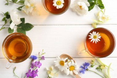 Cam fincanlarda aromatik bitki çayı, farklı çiçekler ve beyaz ahşap masada yeşil yapraklar, düz yatak. Metin için boşluk