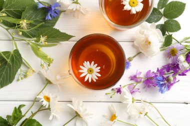 Cam fincanlarda aromatik bitki çayı, farklı çiçekler ve beyaz ahşap masada yeşil yapraklar.