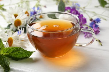 Cam bardakta Aromatik bitki çayı, farklı çiçekler ve beyaz ahşap masada nane şekeri, yakın plan.