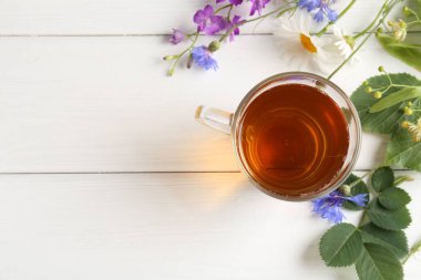 Cam bardakta Aromatik bitki çayı, farklı çiçekler ve beyaz ahşap masa üzerinde yeşil yapraklar, düz yatak. Metin için boşluk