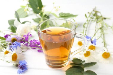 Cam bardakta Aromatik bitki çayı, farklı çiçekler ve beyaz ahşap masada yeşil yapraklar, yakın plan.