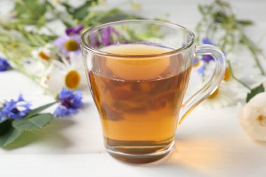 Cam bardakta Aromatik bitki çayı ve beyaz ahşap masada farklı çiçekler, yakın plan.