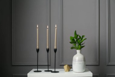 Zarif şamdanlar, yanan mumlar, vazoda dallar ve gri duvarın yanındaki şifoniyerde diğer dekorlar.