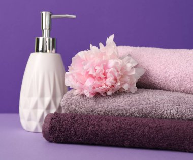 Yumuşak havlu yığını, şakayık çiçeği ve mor arka planda sıvı sabun, yakın plan.