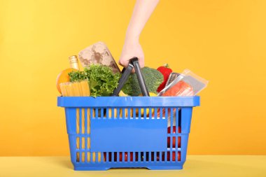 Sarı arka planda ürünlerle dolu alışveriş sepeti olan bir kadın.