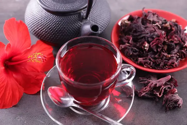 Lezzetli bardakta amber çayı, çaydanlık, kuru roselle sepals ve gri desenli masada güzel çiçek, yakın plan.