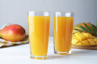 Lezzetli mango suyu bardakta, taze meyve ve yeşil yapraklar beyaz ahşap masada, yakın plan.