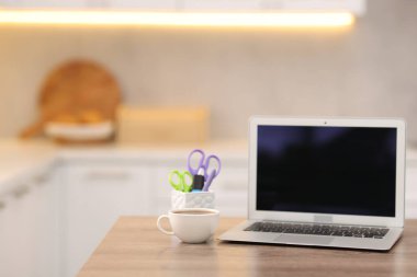 Boş ekranlı dizüstü bilgisayar, mutfaktaki ahşap masada bir fincan sıcak içecek ve kırtasiye malzemesi. Tasarım için model