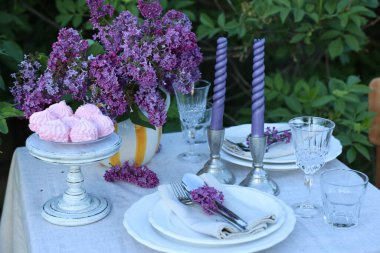 Güzel leylak çiçekleri, zephyrler, tabaklar, çatal bıçaklar, bardaklar ve mumlar bahçede.