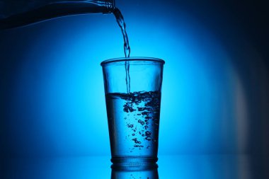 Mavi arkaplanda şişeden cama su dökmek. Arka ışıklandırma