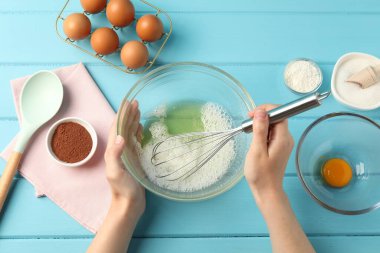 Bir kadın açık mavi ahşap masada kasede yumurta akı çırpıyor.