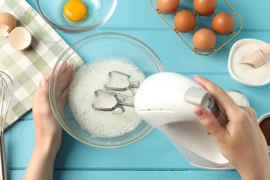 Bir kadın açık mavi ahşap masada el çırpıcı ile yumurta beyazı çırpıyor.