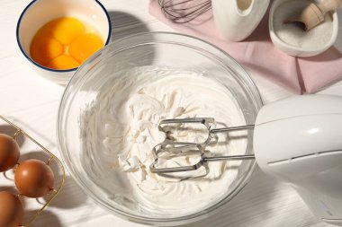 Beyaz ahşap masada çırpılmış krema, yumurta ve diğer ürünlerle el çırpma makinesi.