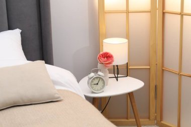 Yan masada lamba, çalar saat ve yatağın yanında çiçek var.