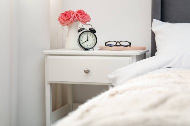Yatağın yanında çalar saati, çiçekleri ve bardakları olan bir komodin.