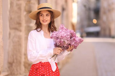 Şapkalı gülümseyen kadın, bina yakınlarında bir demet leylak çiçeğiyle.