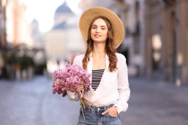 Şapkalı gülümseyen kadın, şehir caddesinde bir demet leylak çiçeğiyle.