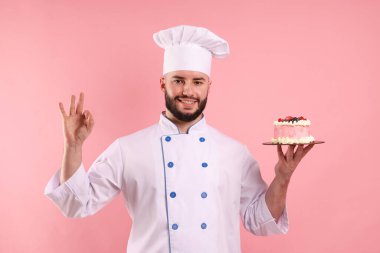 Üniformalı mutlu pastacı elinde böğürtlen ve pembe arka planda iyi bir jest yapıyor.