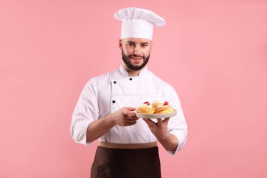 Üniformalı mutlu pastacı elinde pembe arka planda çilekli profiterol tutuyor.