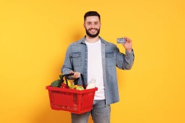 Turuncu arka planda ürünlerle dolu alışveriş sepeti ve kredi kartı olan bir adam.