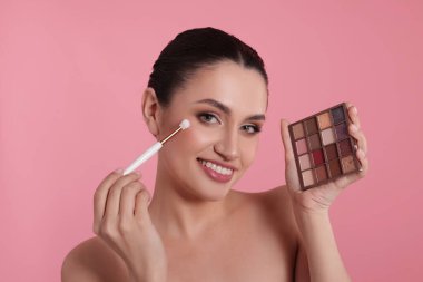 Göz farı paleti ve fırçasıyla gülümseyen kadın pembe arka plana makyaj yapıyor.
