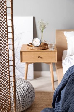 Komodinin yanında çalar saat, vazoda çiçekler ve yatağın yanında bir bardak su.
