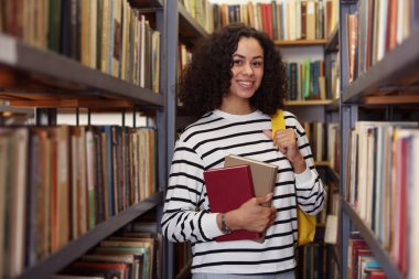 Halk kütüphanesinde rafların yanında kitapları ve sırt çantaları olan bir kadın.