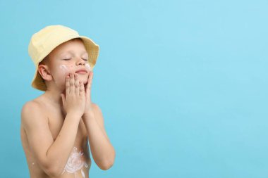 Küçük çocuk açık mavi arka planda cildine güneş kremi sürüyor.