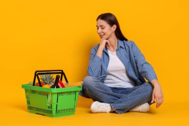 Gülümseyen kadın ve alışveriş sepeti sarı arka planda ürünlerle dolu.