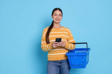 Açık mavi arka planda akıllı telefonu ve sepeti olan gülümseyen bir kadın. İnternetten alışveriş