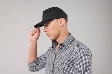 Genç adam açık gri arka planda şık bir siyah beyzbol şapkası giyiyor.