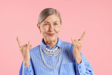 Üst düzey kadın zarif inci takılar takıyor ve pembe arka planda taş jestler yapıyor.