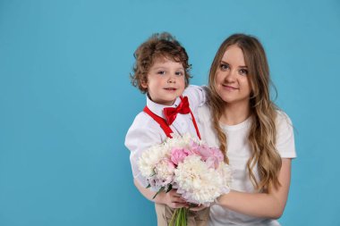 Küçük çocuk annesine açık mavi arka planda şakayık çiçekleri veriyor.