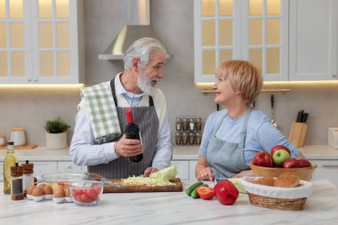 Usta adam mutfakta yemek pişirirken karısına şarap öneriyor.