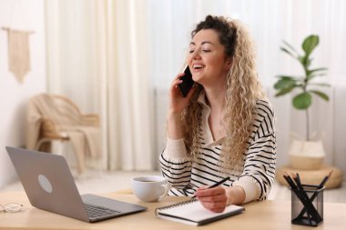 Gülümseyen kadın akıllı telefonla konuşuyor ve ofis masasında çalışıyor.