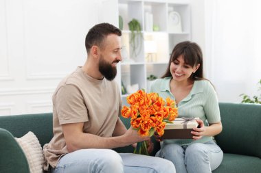 Bir adam evdeki kanepede sevgili karısına güzel laleler sunuyor.