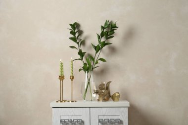 Zarif şamdanlar, yanan mumlar, vazoda dallar ve bej duvarın yanındaki şifoniyerde diğer dekorlar.