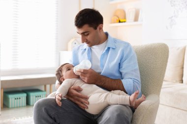 Baba küçük bebeğini evdeki koltukta şişeyle besliyor.