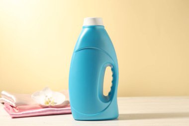 Bir şişe yumuşatıcı, temiz kıyafetler ve açık sarı arka plana karşı beyaz ahşap masa üzerinde orkide çiçeği.