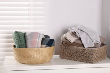 Beyaz komodinin üstündeki sepetlerde yün kazaklar ve eşarplar.