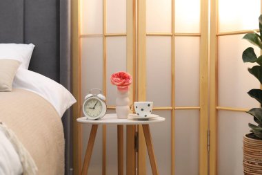 Çalar saati olan yan masa, bir fincan çay ve yatağın yanında çiçek.