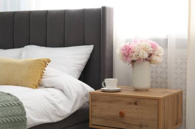 Yatağın yanında kahve ve çiçek olan bir komodin.